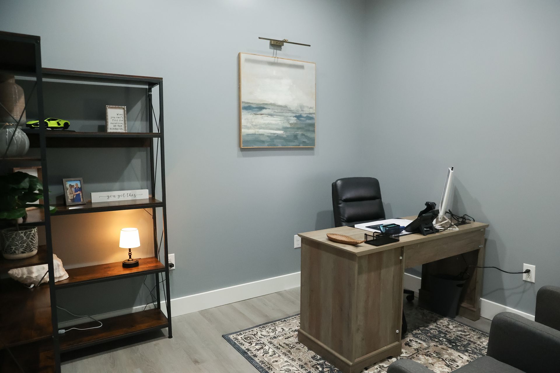 Office with desk, shelving, art, and chair against gray walls and light floor.