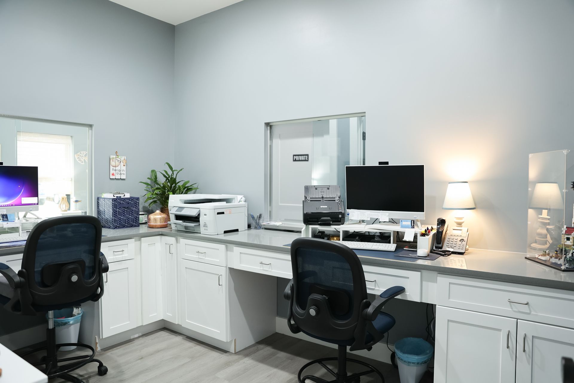 Office with two desks, computers, printers, chairs, and white cabinets. Walls are gray.