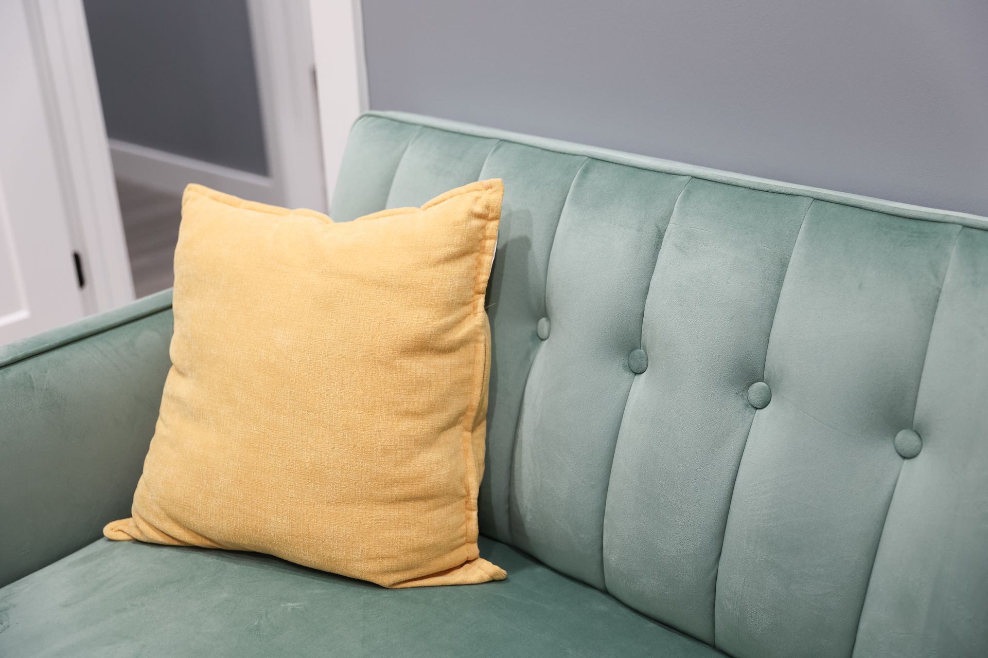 A yellow pillow rests on a teal velvet couch with button tufting, against a gray wall.
