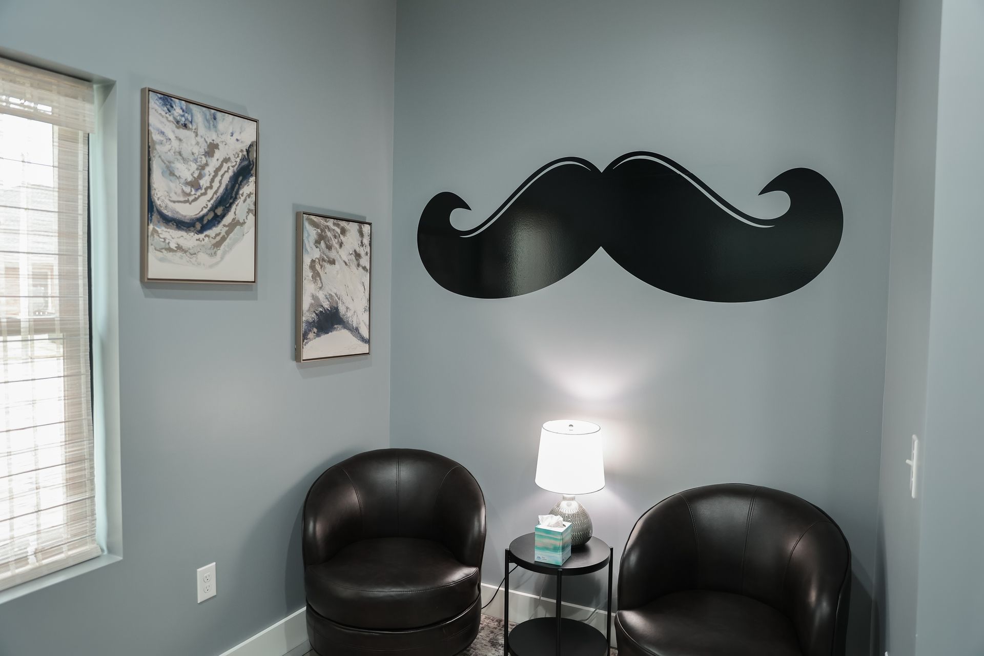 A waiting room with two black chairs, a table with a lamp, and a large mustache decal on a blue wall.