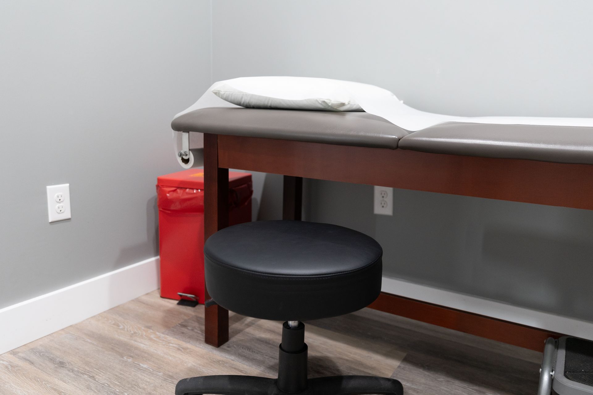 A medical exam room with an examination table, stool, and a red trash can.