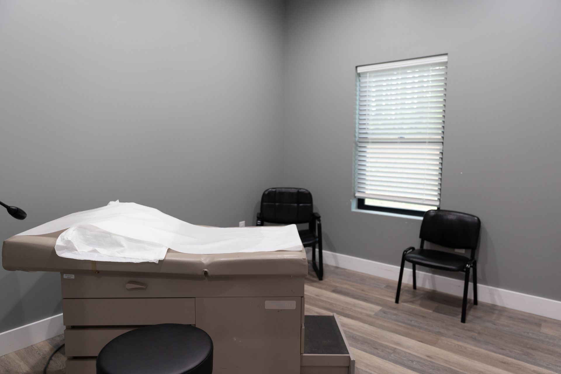 Examination room: gray walls, medical table, chairs, window with blinds.