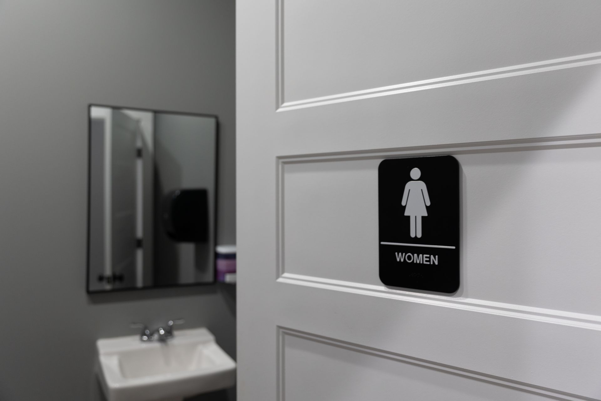 A women's restroom door with a black sign and a female symbol. The interior features a sink and mirror.