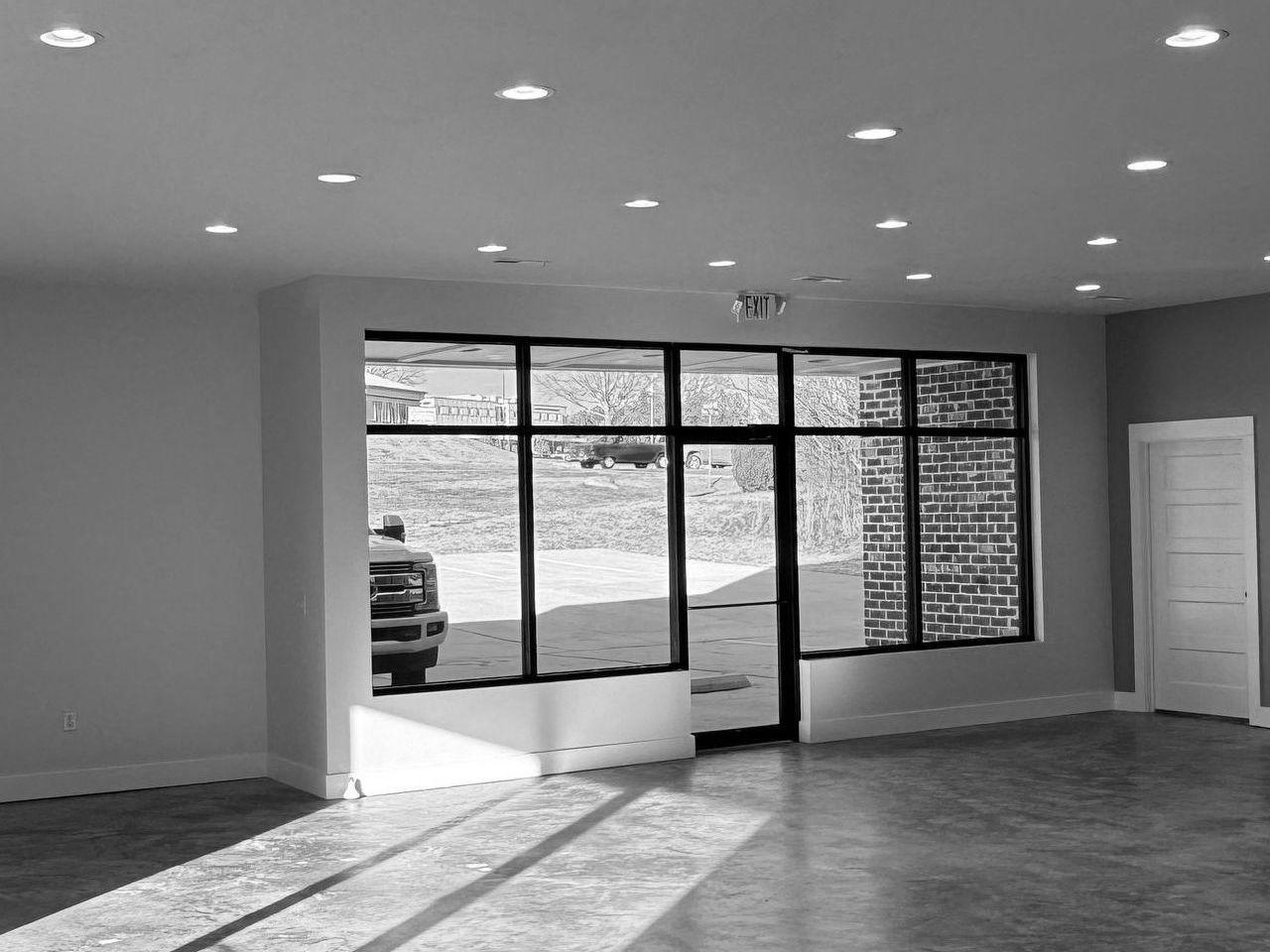 Empty, gray interior with large windows and a brick accent wall; a car is visible outside.