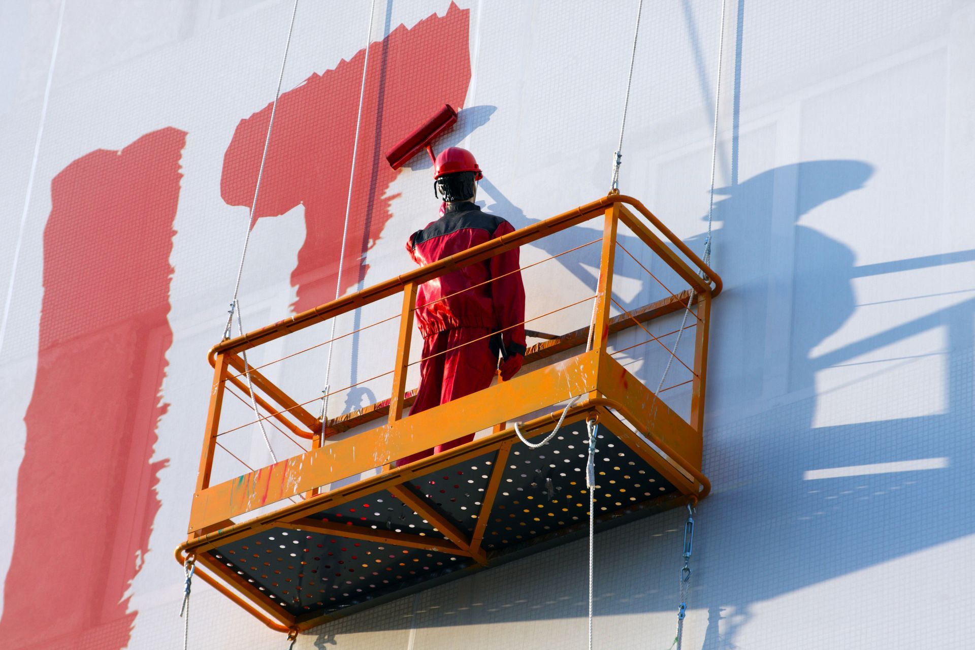 Worker in red overalls on an orange platform painting red letters on a white wall.