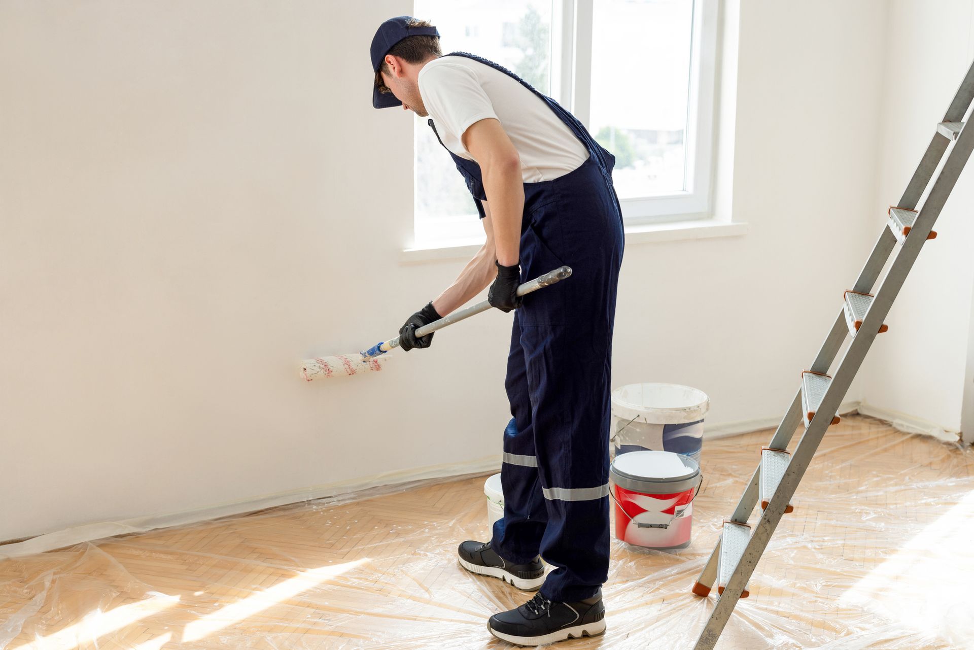 Painter in navy overalls rolls white paint on a wall; paint cans and ladder in a room.