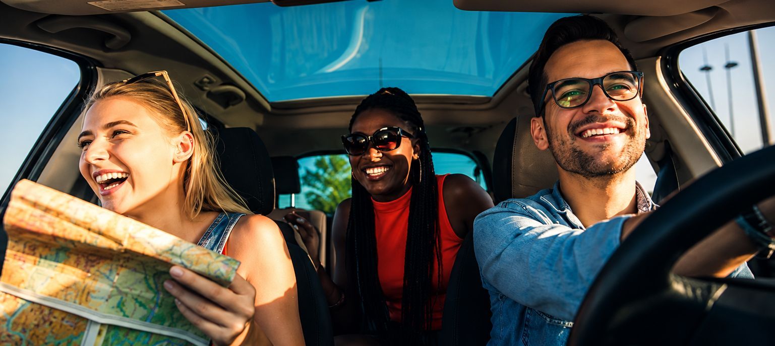Three friends smile in a car, one holding a map on a sunny road trip