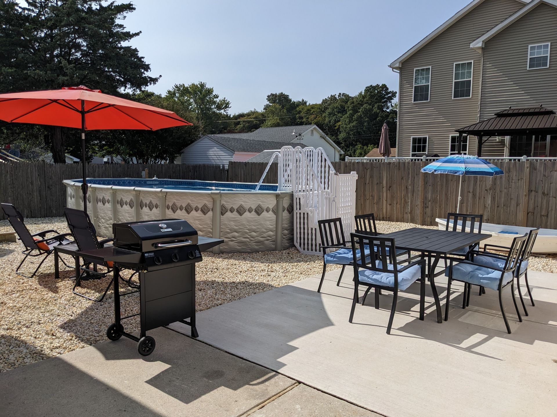 A patio with a table , chairs , grill and umbrellas in front of a pool.