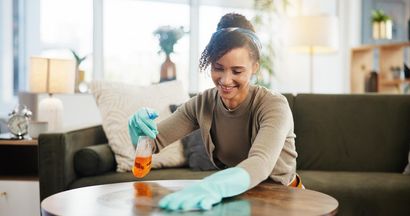 Woman wearing gloves cleaning a wooden table in a living room, smiling.