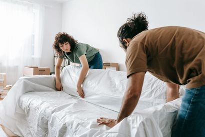 Two people covering a sofa with white plastic in a room with boxes, preparing for a move.