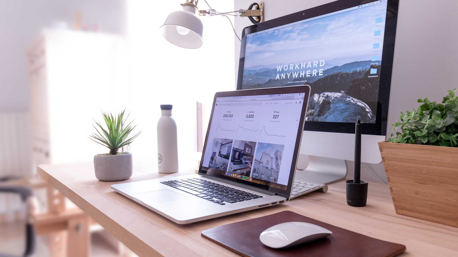 Minimalist desk setup with laptop, monitor, lamp, plant, water bottle, and mouse in a bright office.