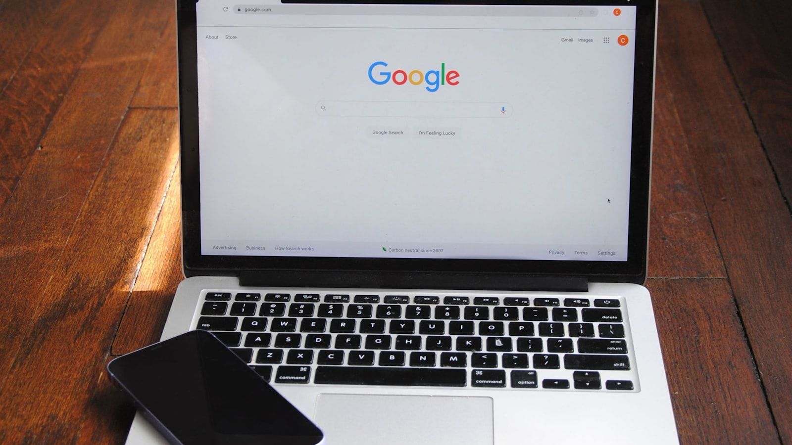 Laptop on wooden table displaying the Google homepage, with a black phone beside it.