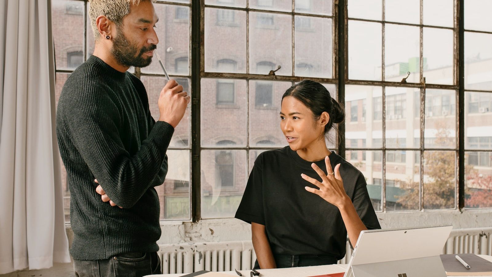 Two people talking in a bright office by large windows, one standing and one seated at a desk.