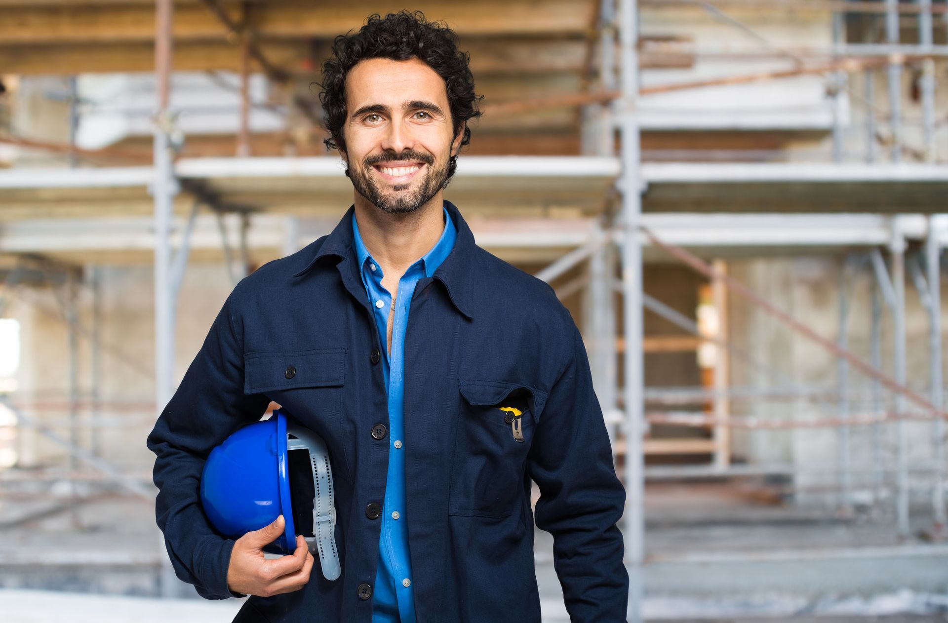 Smiling worker holding a blue hard hat in a construction site with scaffolding behind him