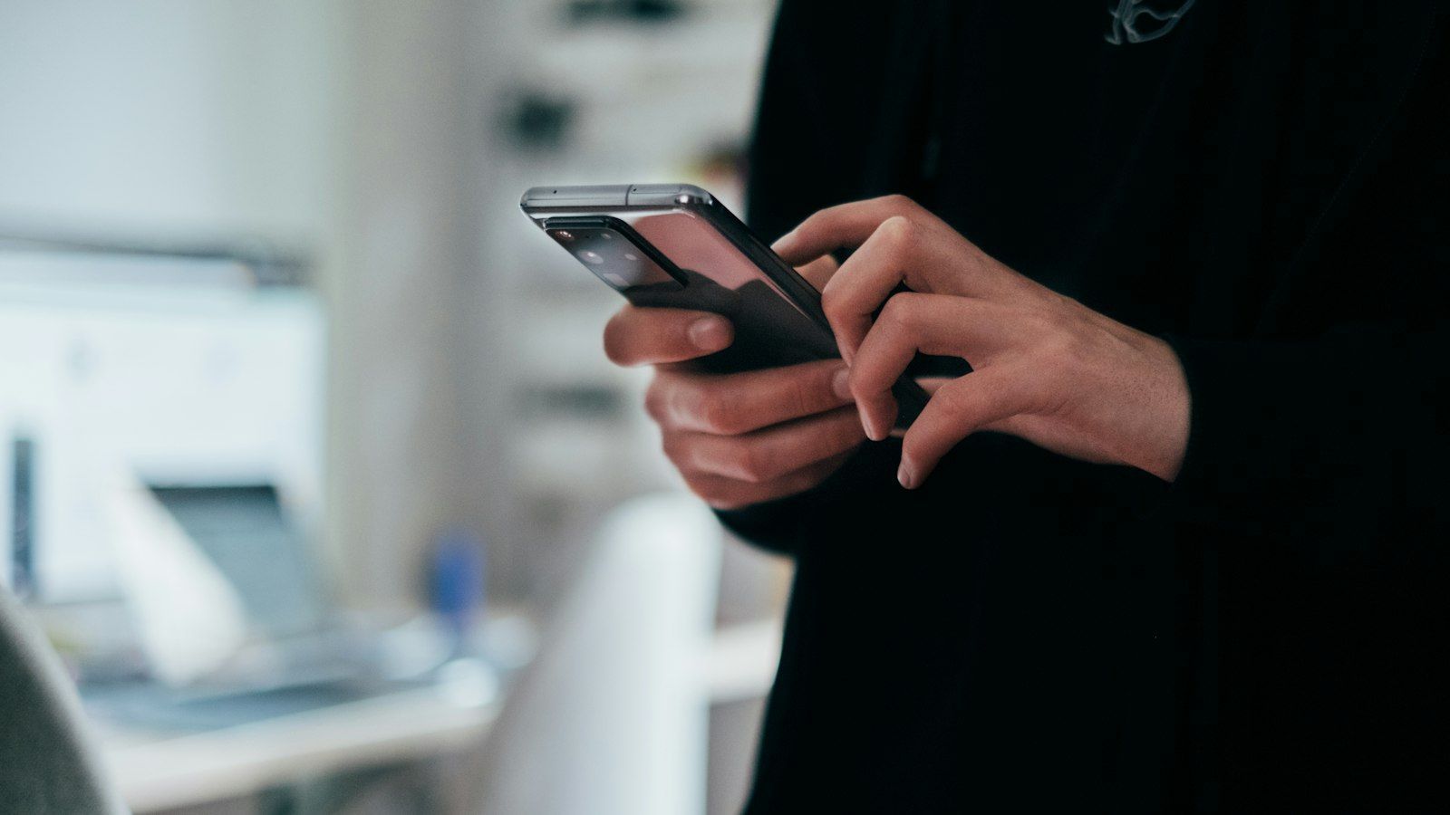 Person in black clothing using a smartphone indoors, with a blurred desk and laptop in the background