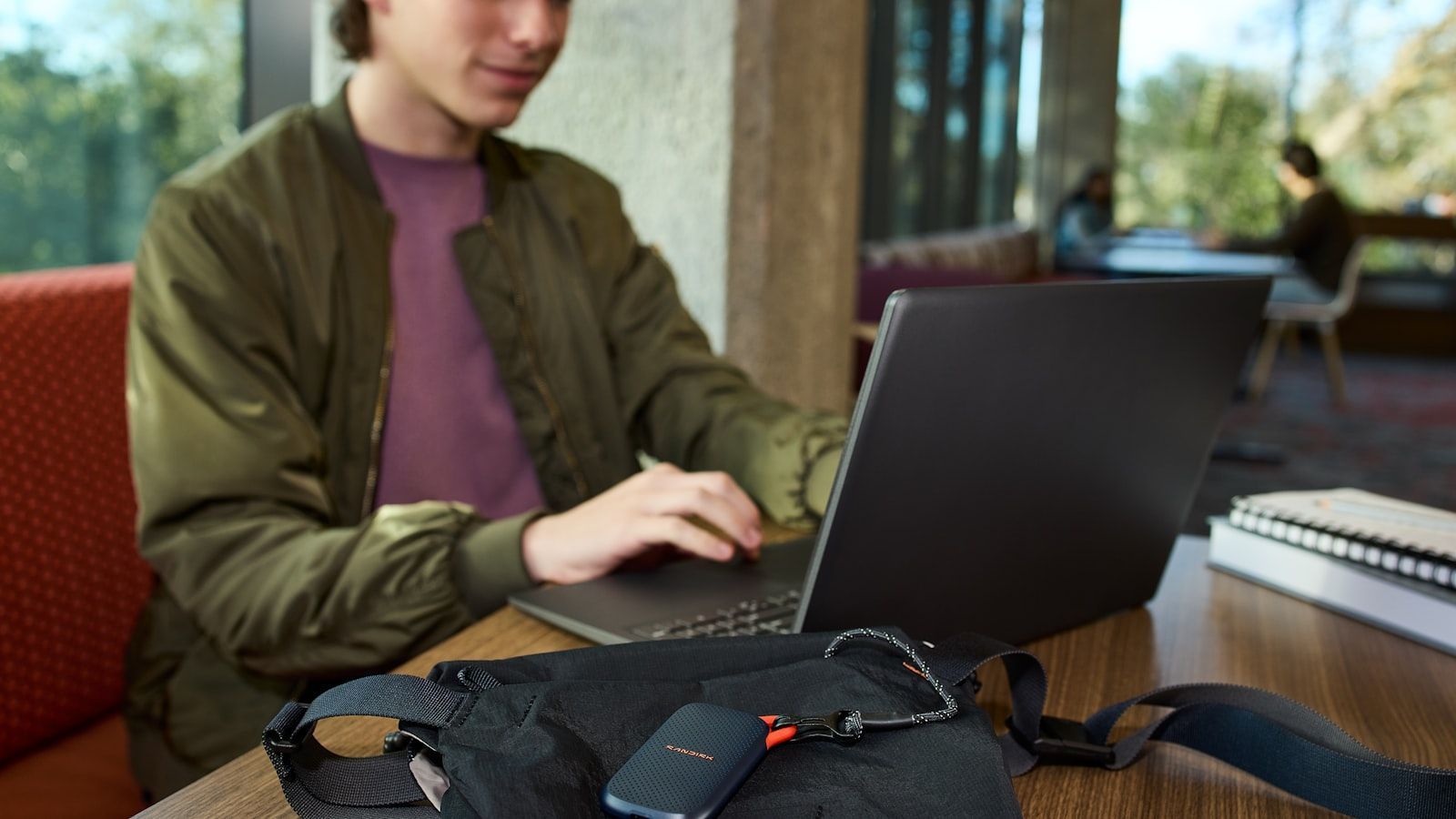 Person using a laptop at a table with a black bag in front, indoors near a window