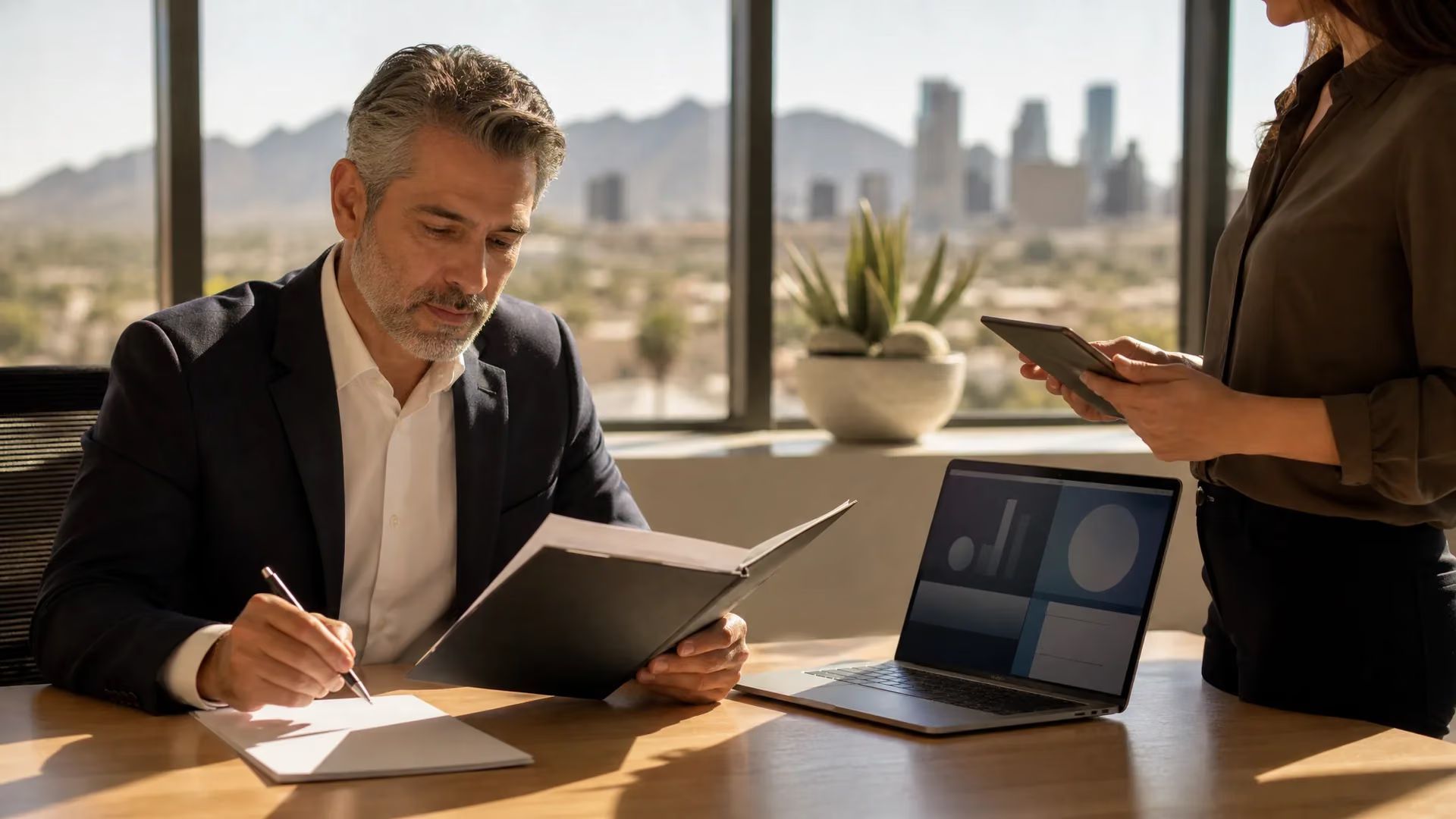 Professional reviewing documents at a sunlit office desk while a colleague stands nearby with a tablet.