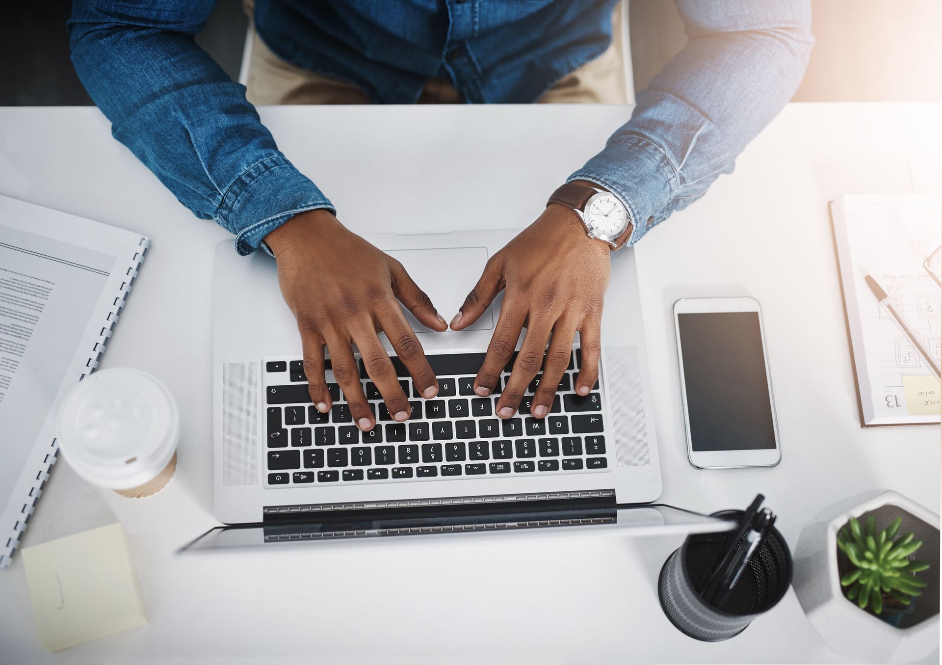 Hands typing on a laptop at a white desk with coffee, phone, notebook, and plant
