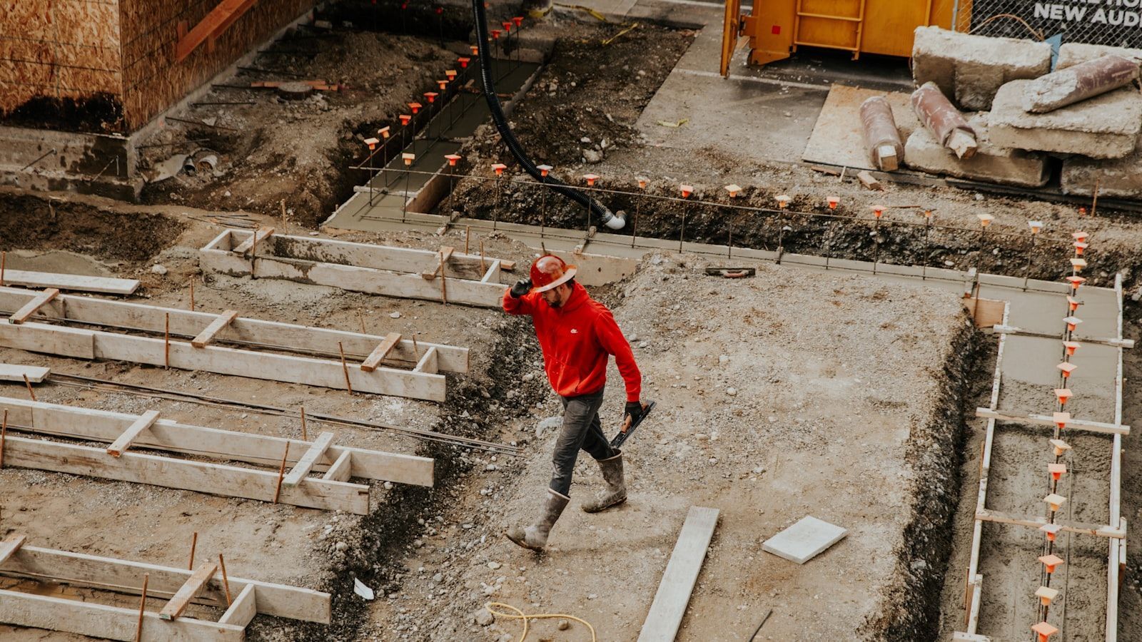 Worker in red jacket walking across a muddy construction site with wooden forms and concrete slabs