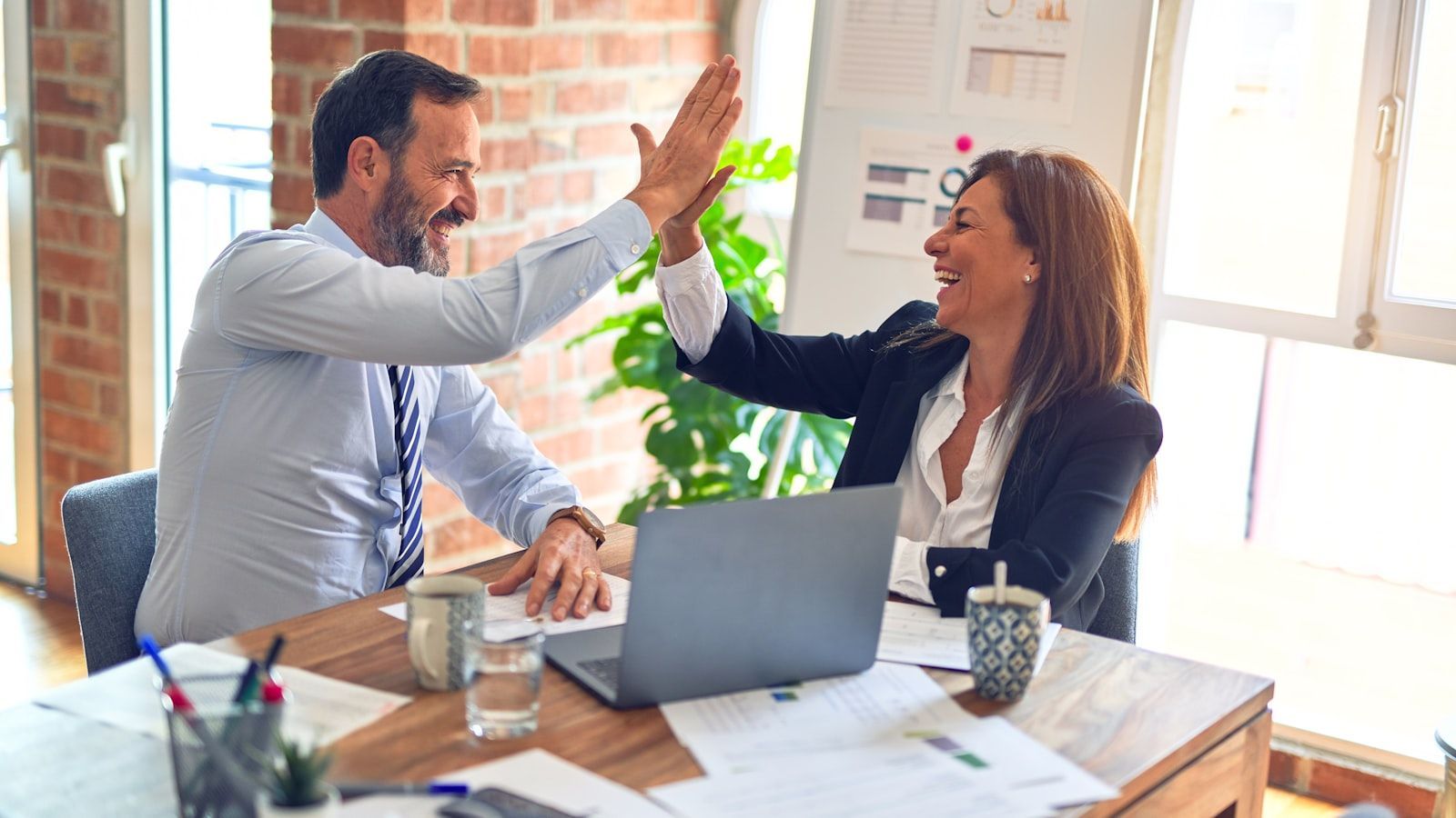 Two coworkers high-five at a laptop in a bright office with brick walls and large windows.