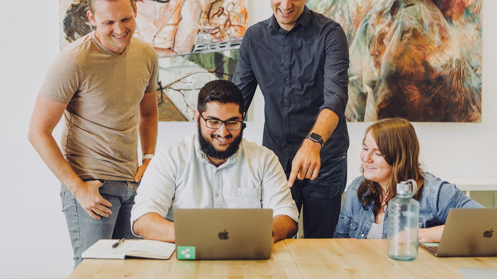 Four coworkers smiling around a laptop in a bright office meeting room