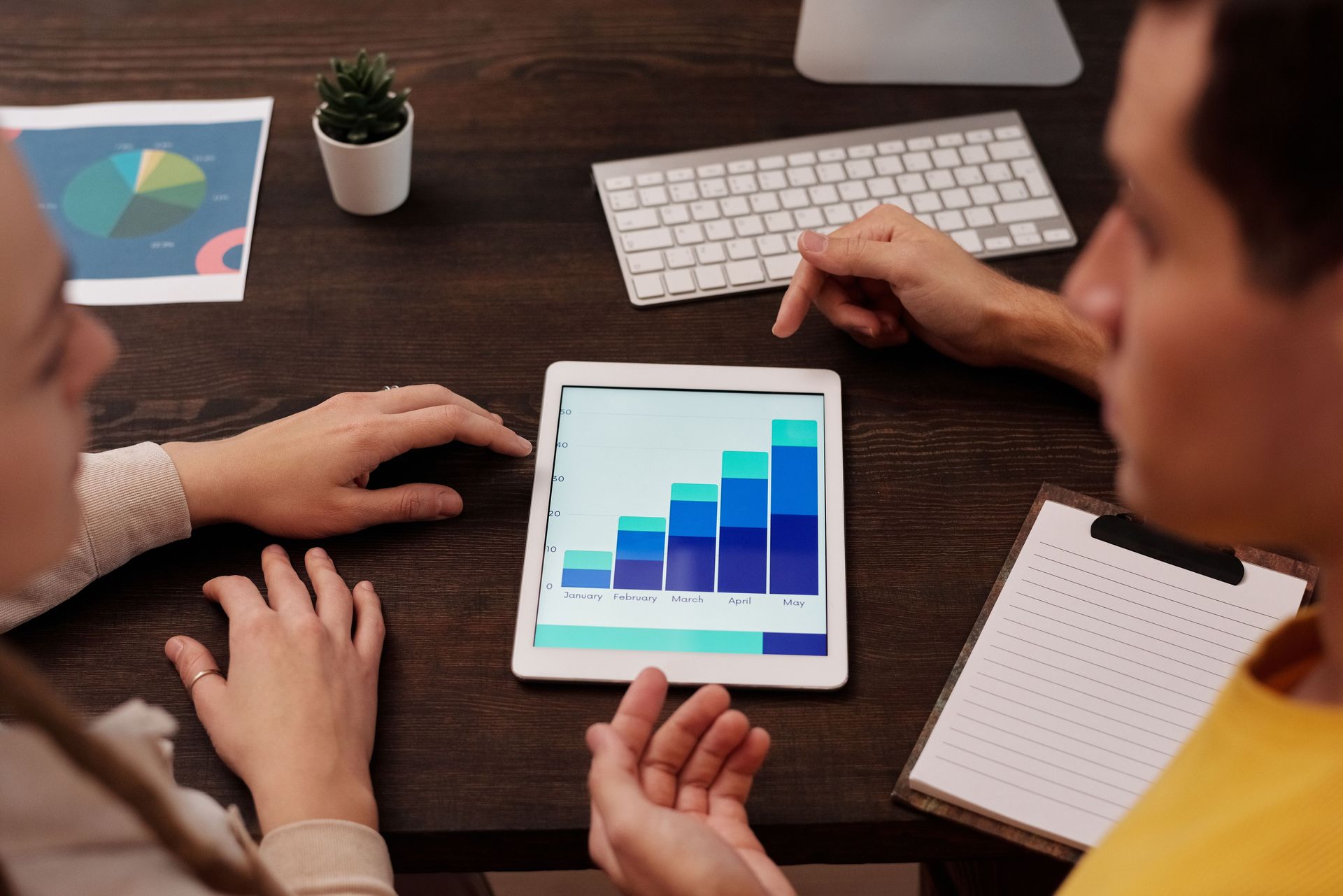 Team meeting around a tablet showing a blue bar chart on a desk with a keyboard and notebook