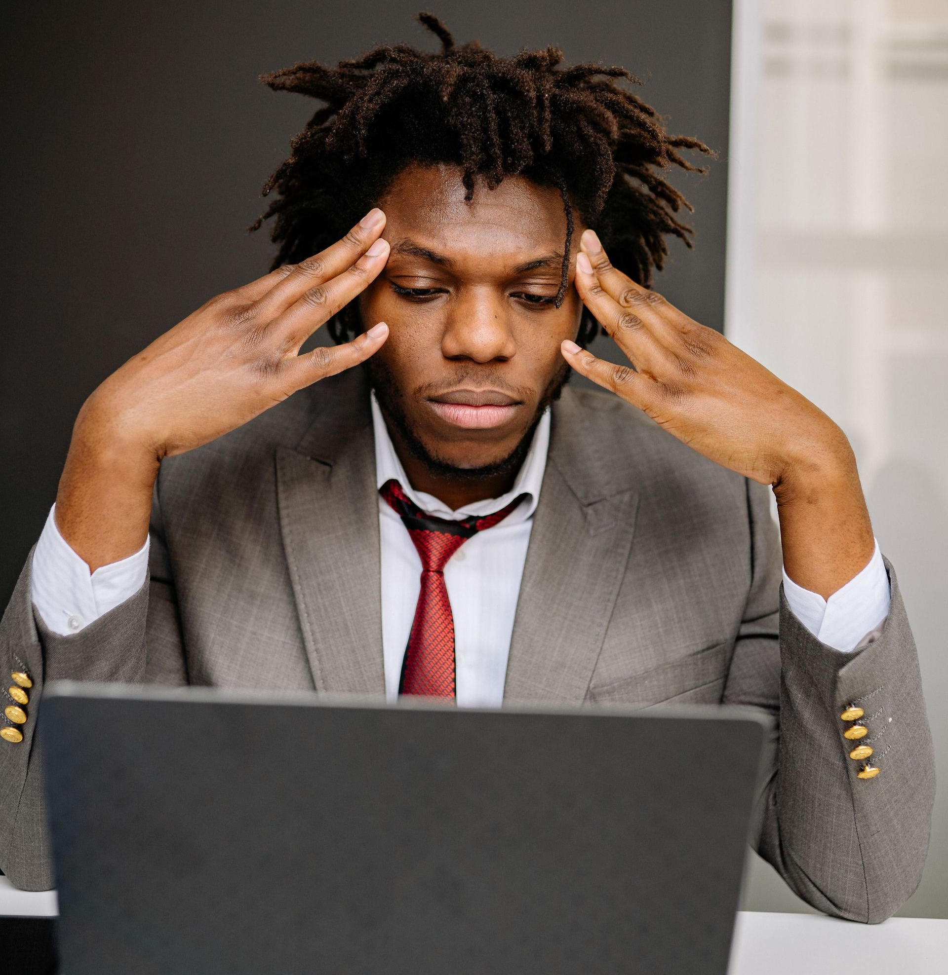 Man in a gray suit rubbing his temples at a laptop, looking stressed in an office setting