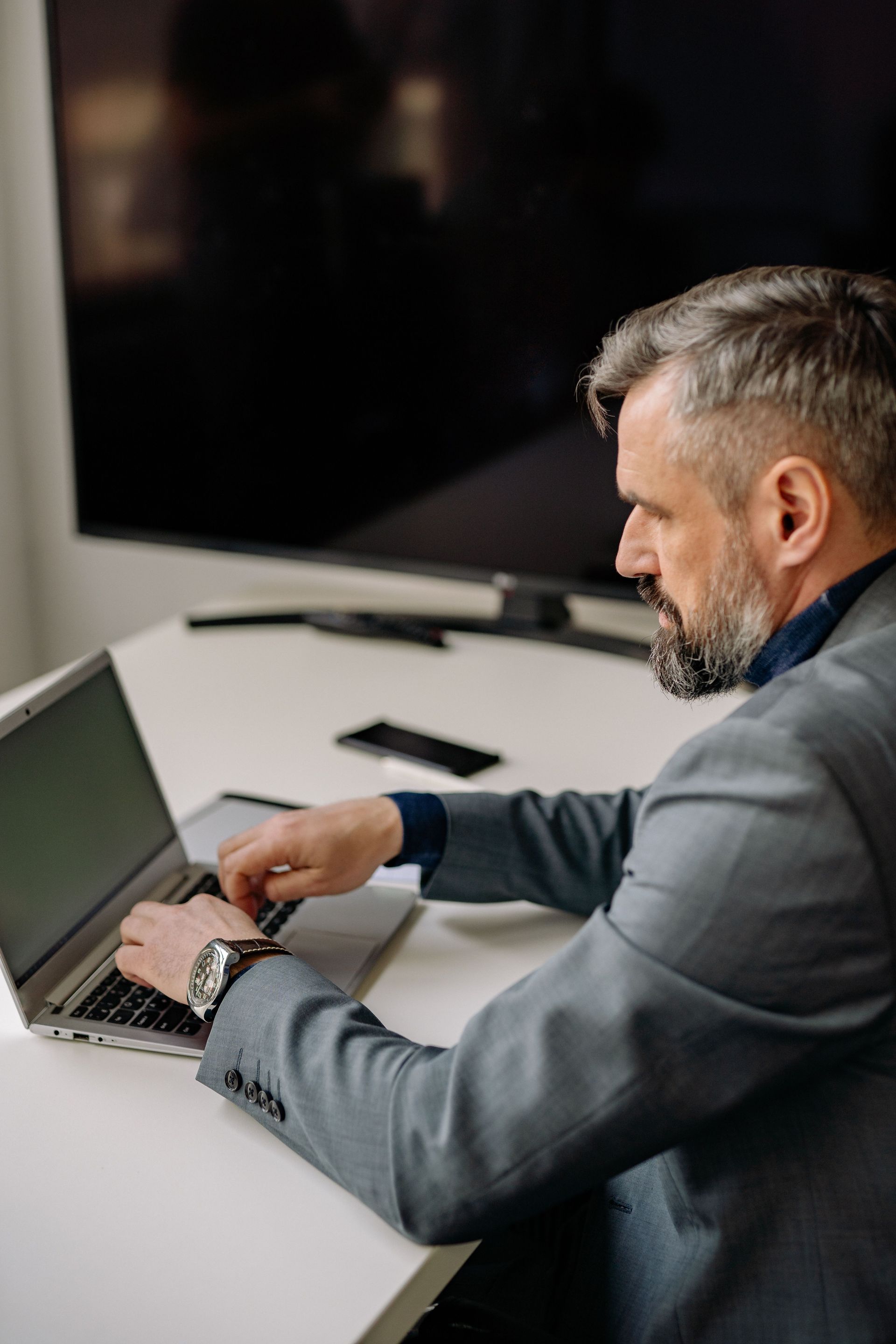 Man typing on a laptop at a desk in a modern office, with a large monitor in the background