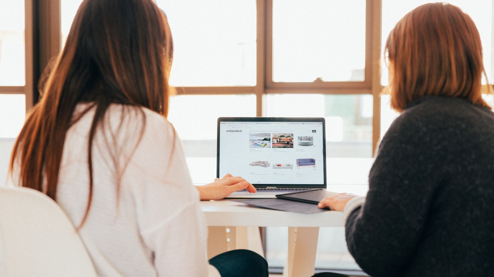 Two people sit at a table facing a laptop in a bright room with large windows.