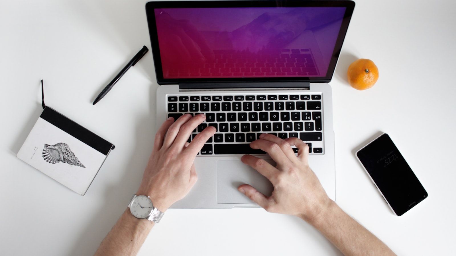 Hands typing on a laptop at a white desk with a phone, notebook, pen, and orange nearby