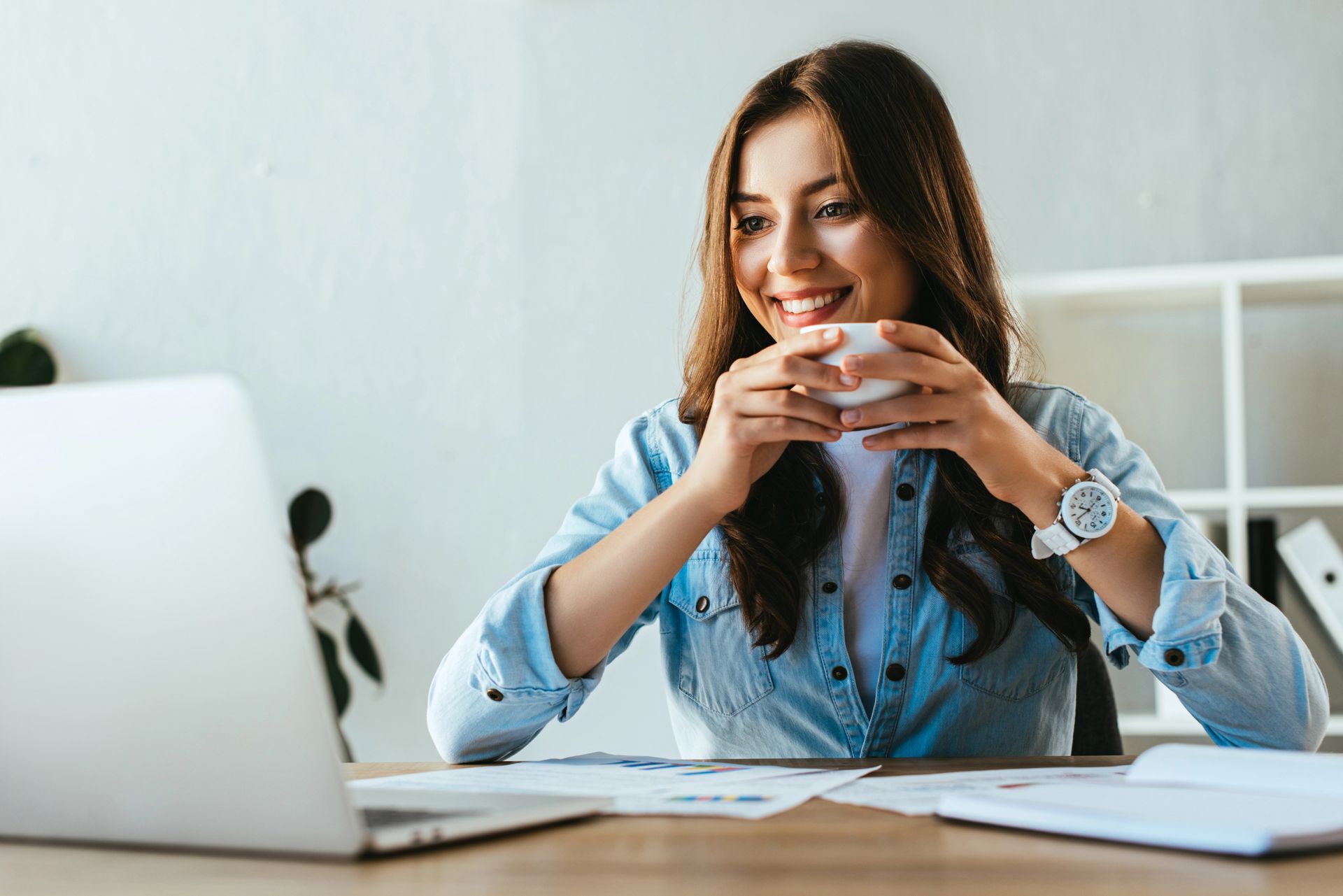 Woman smiling at a desk, holding a cup beside a laptop and open notebook in a bright office