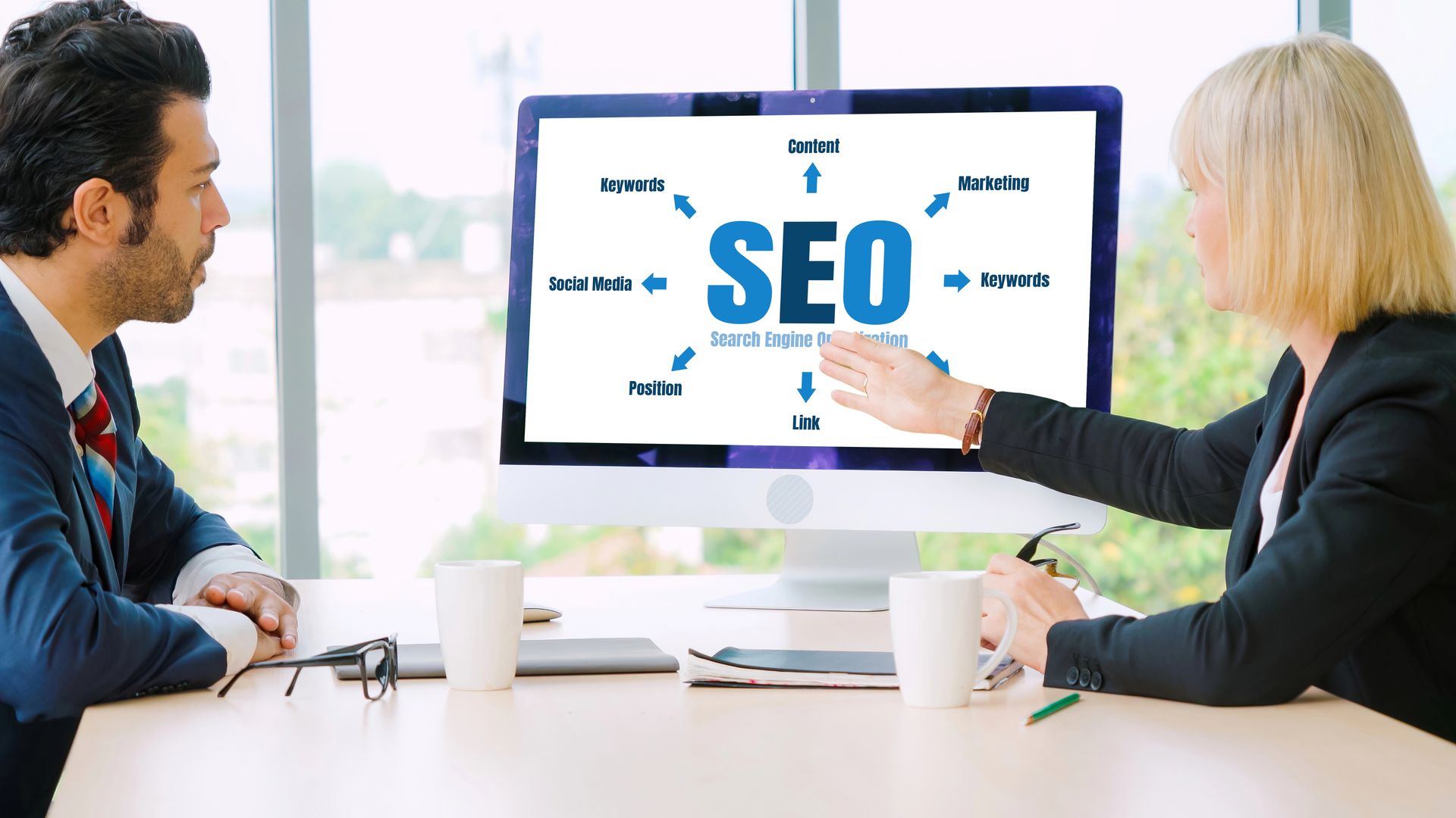 Two people in a meeting pointing at a monitor displaying “SEO” with marketing keywords around it