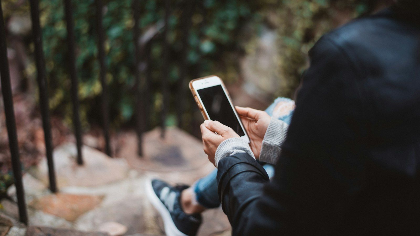 Person holding a smartphone while seated outdoors near a fence and trees.