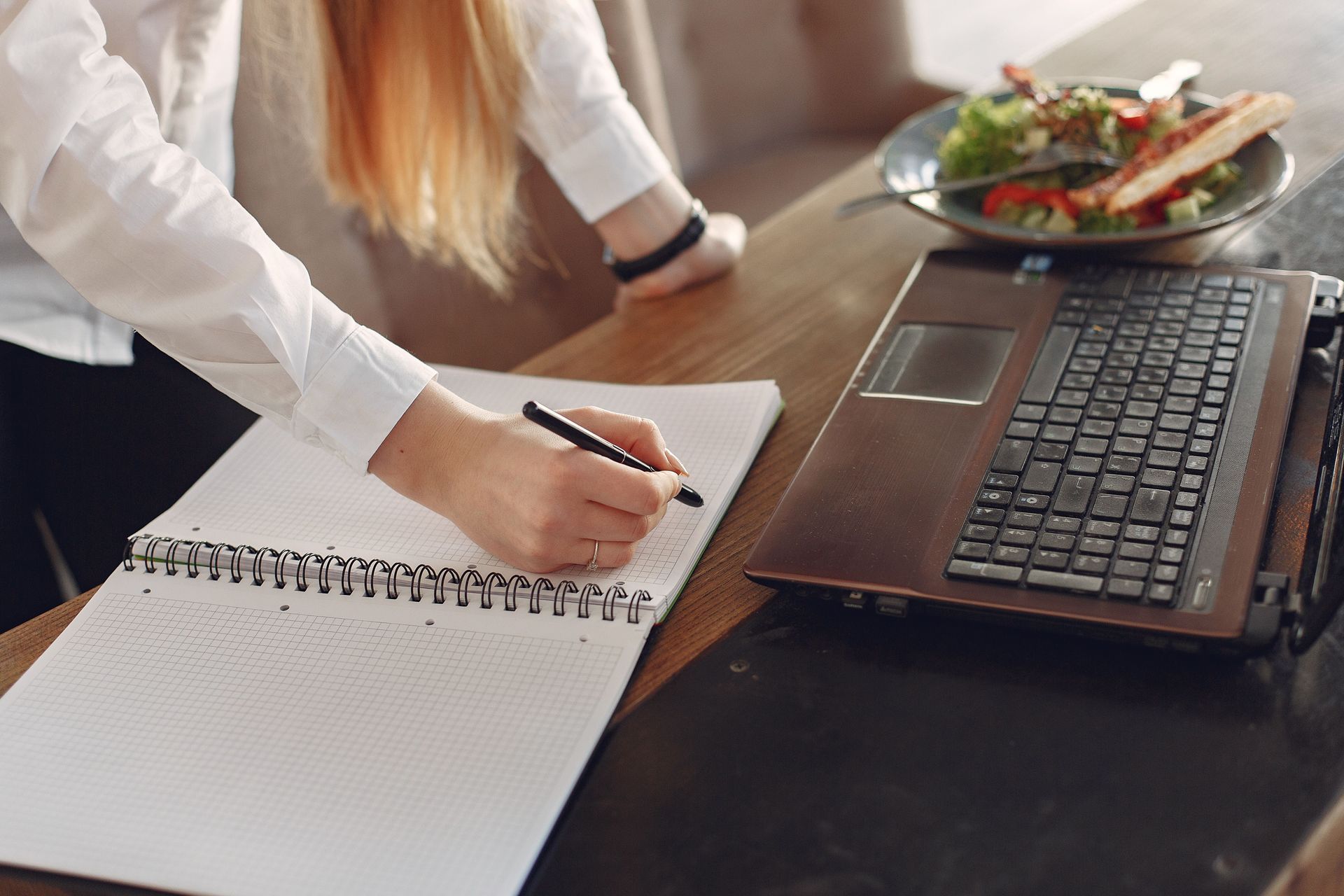 Person writing in a spiral notebook beside an open laptop on a desk with a plate of food