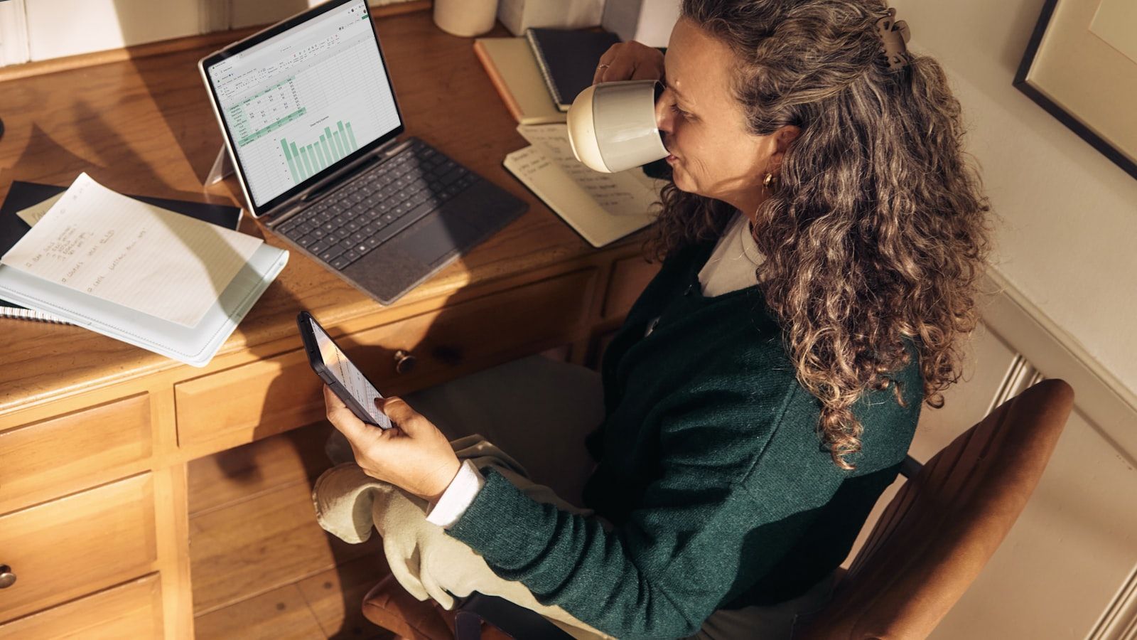 Woman working at a wooden desk with a laptop and tablet in a sunlit room