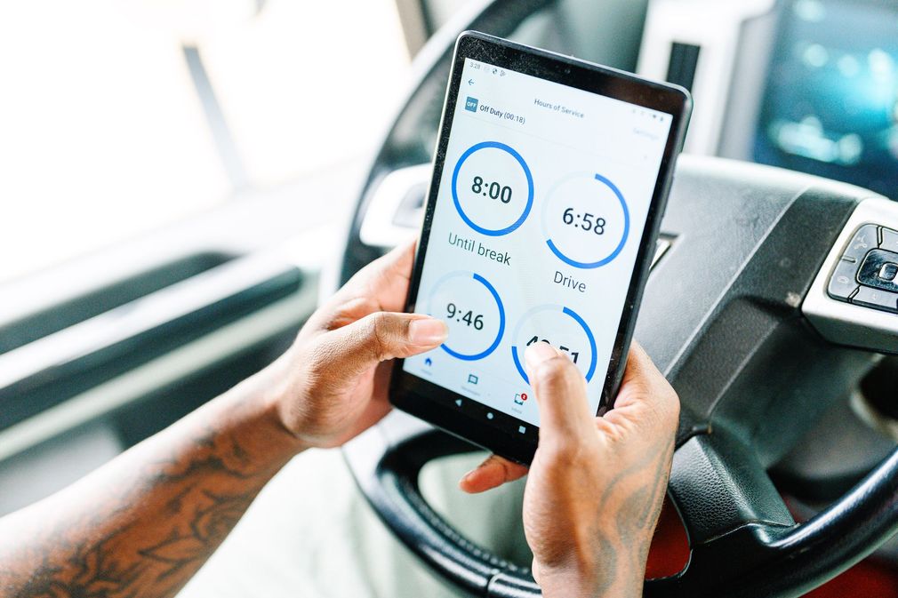 Hands hold a tablet displaying an electronic logbook while positioned over a truck steering wheel.