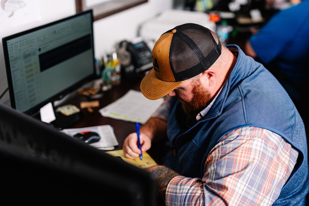A bearded person in a baseball cap and plaid shirt sits at a desk, writing on a notepad while facing a computer monitor.