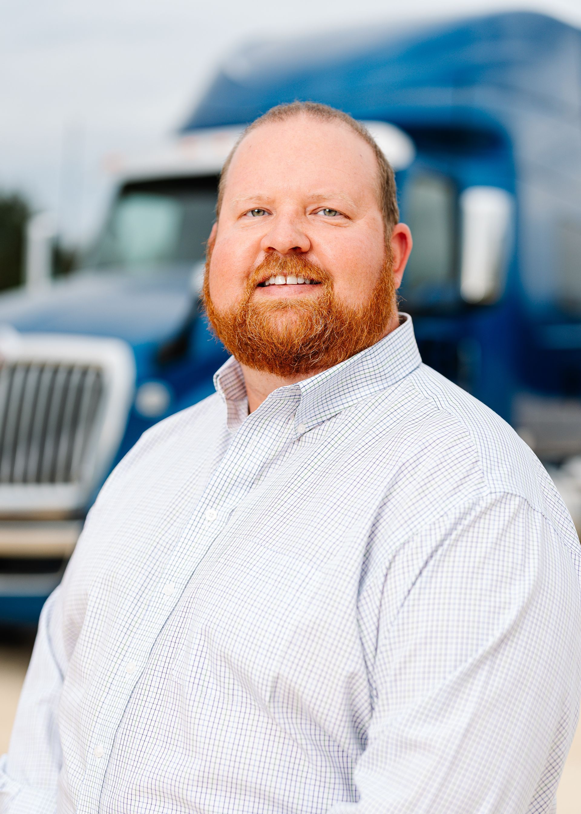 A smiling person with a beard, wearing a light patterned button-down shirt, stands in front of a blue semi-truck.