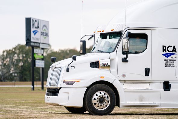 A white RCA branded semi-truck parked in a field with a business sign in the background.