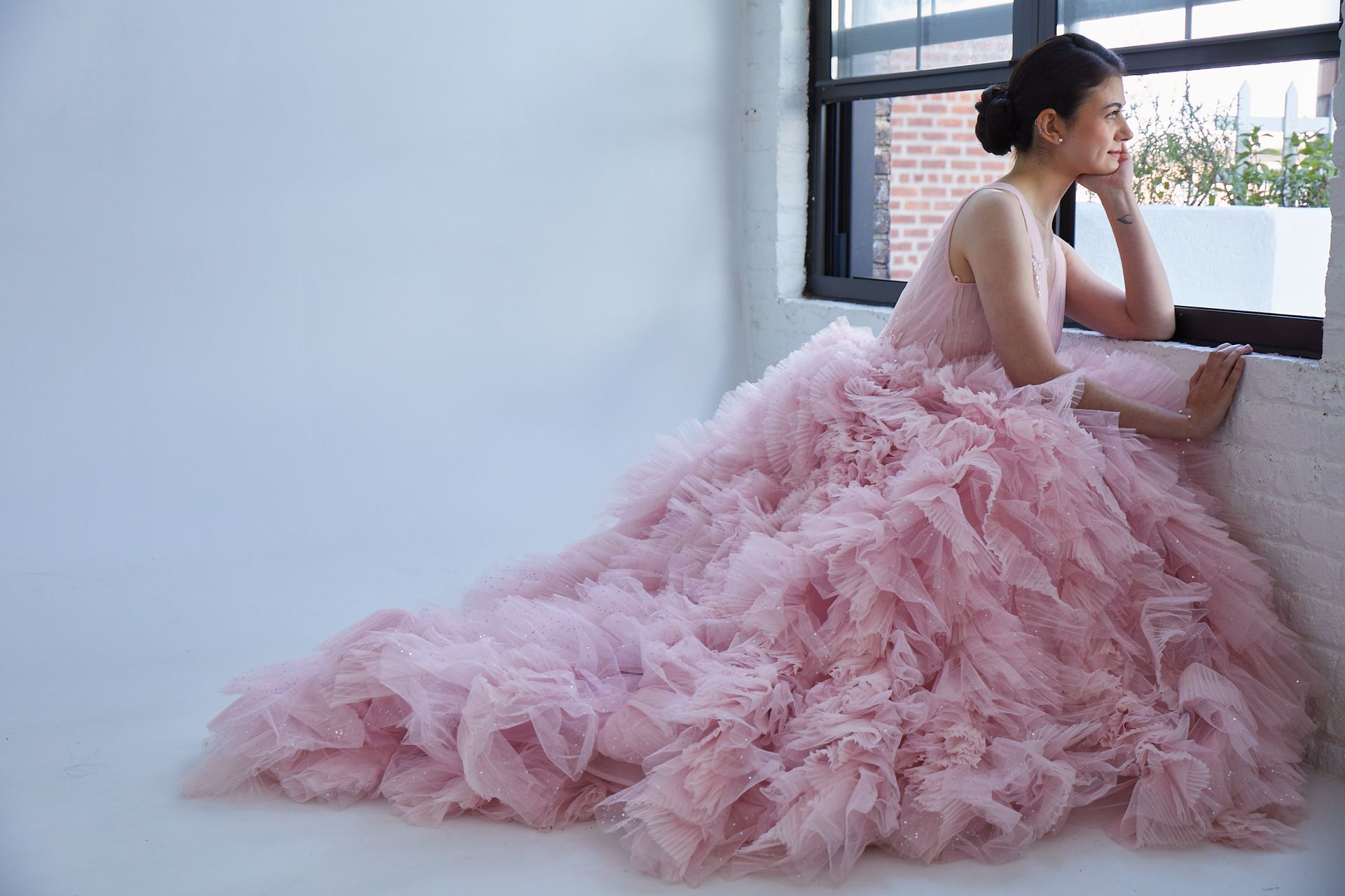 Woman in a pink tulle gown sitting by a window, gazing outside.