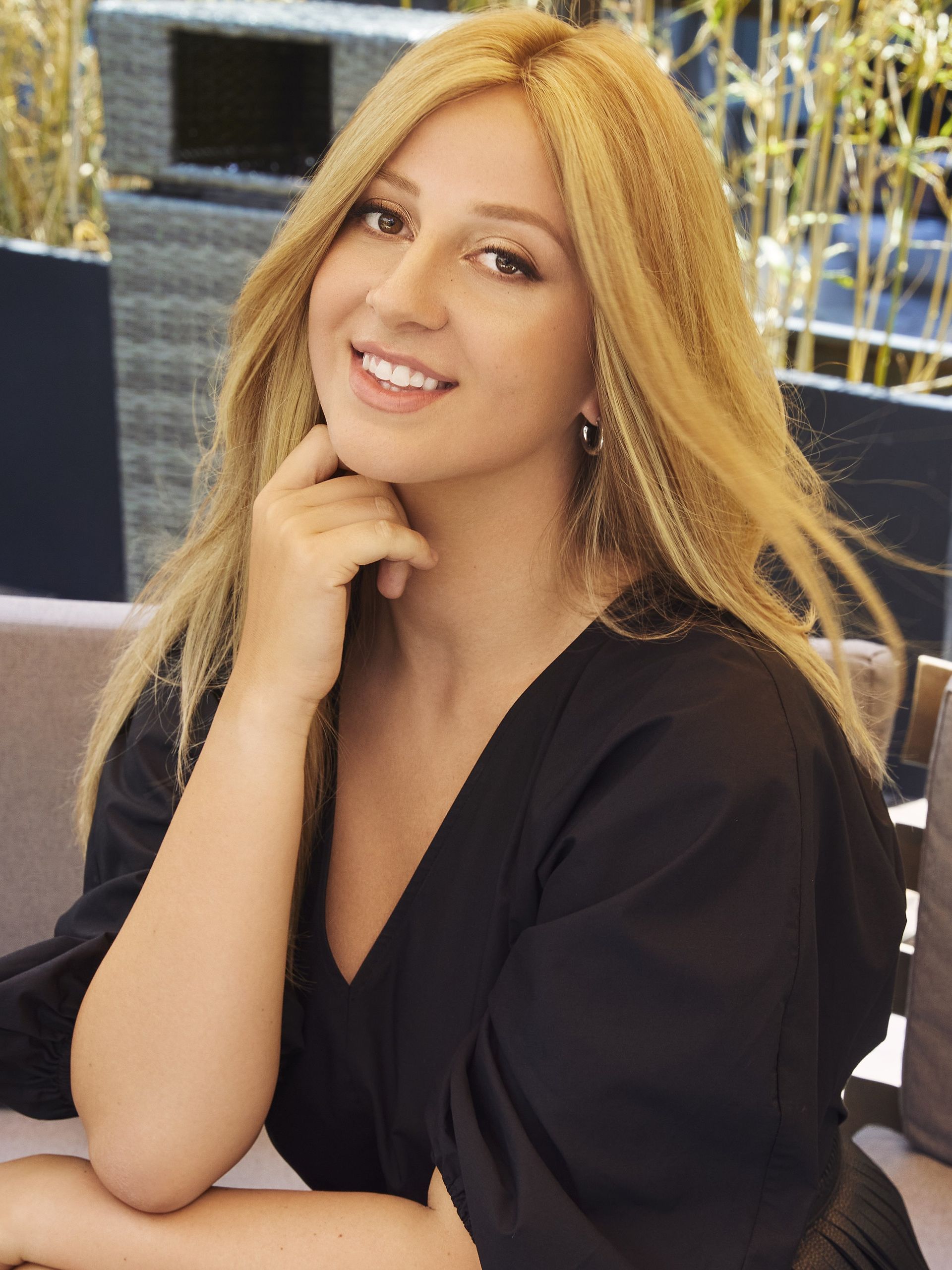 Smiling woman with long blonde hair in a black top, resting her chin on her hand at an outdoor table.
