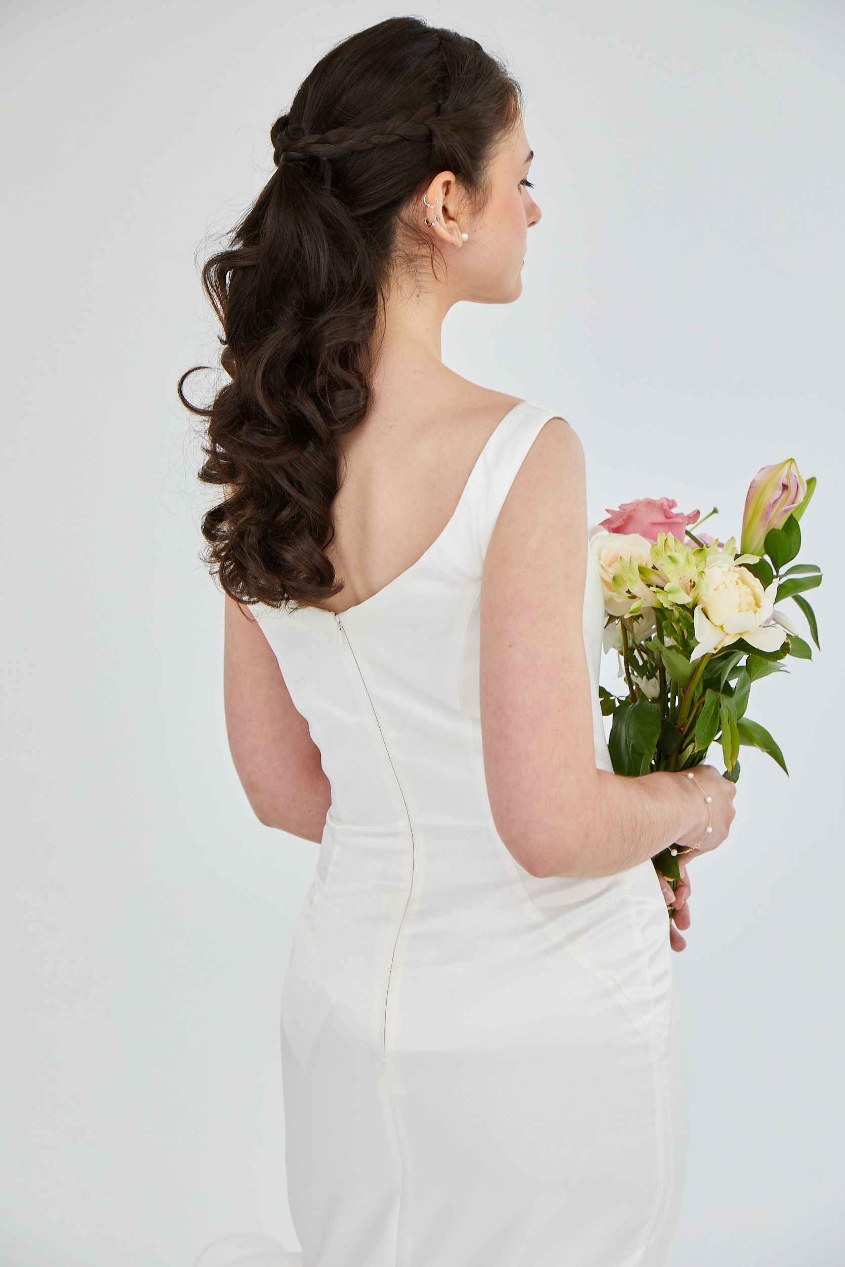 Woman in a white dress holding a bouquet of flowers, seen from behind against a plain background.