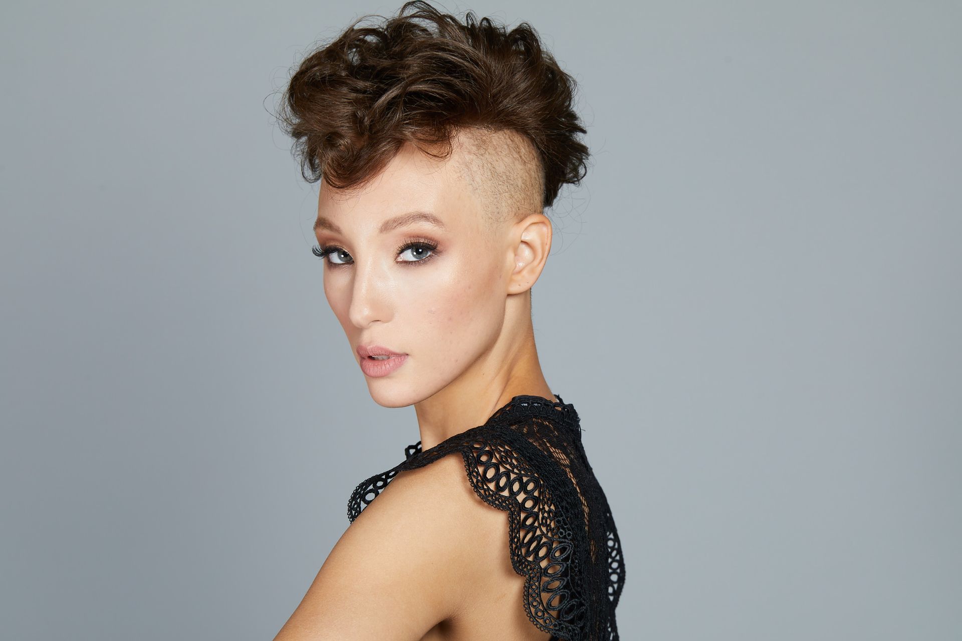 Portrait of a person with a curly undercut hairstyle and black lace top against a gray background