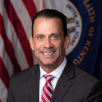A man in a suit and tie is smiling in front of a flag that says kentucky