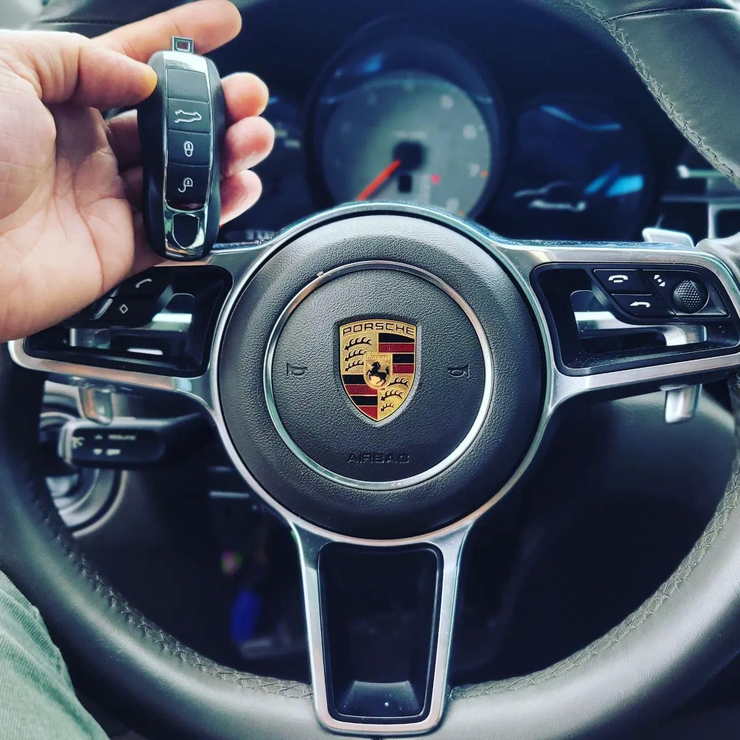 Hand holding Porsche car key in front of the steering wheel with the Porsche logo.