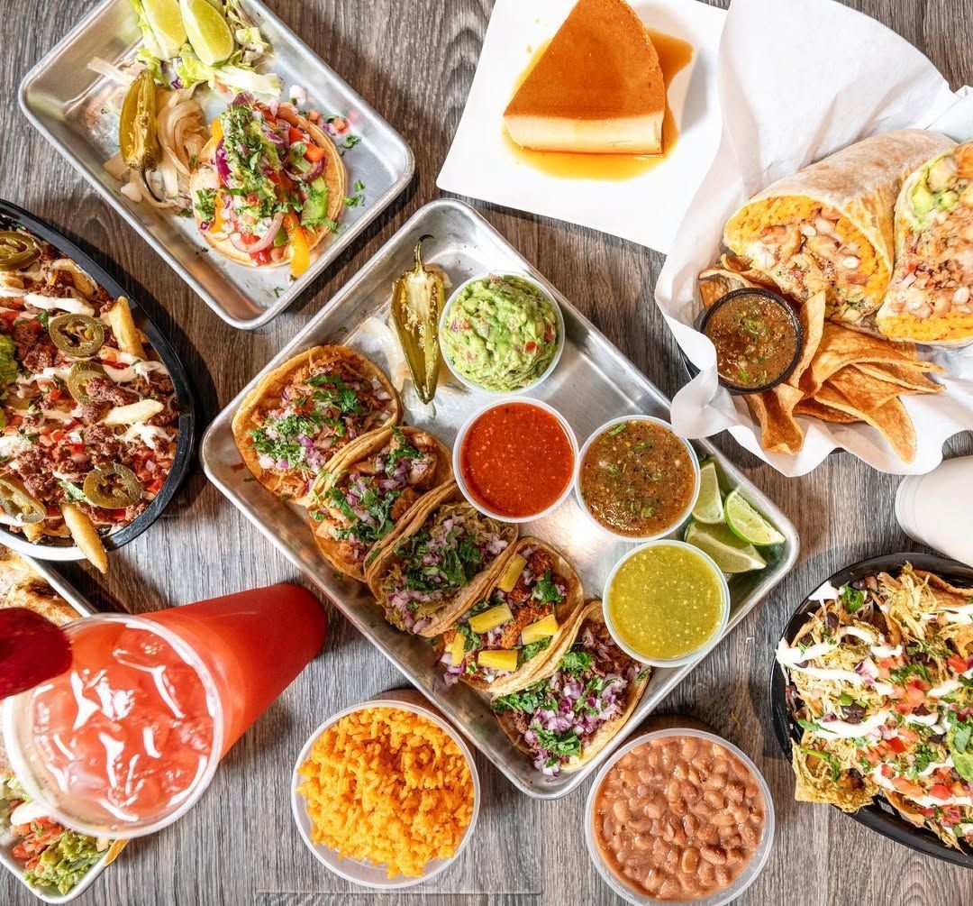Assorted colorful street foods in trays and bowls, including tacos, sauces, and snacks on a wooden table