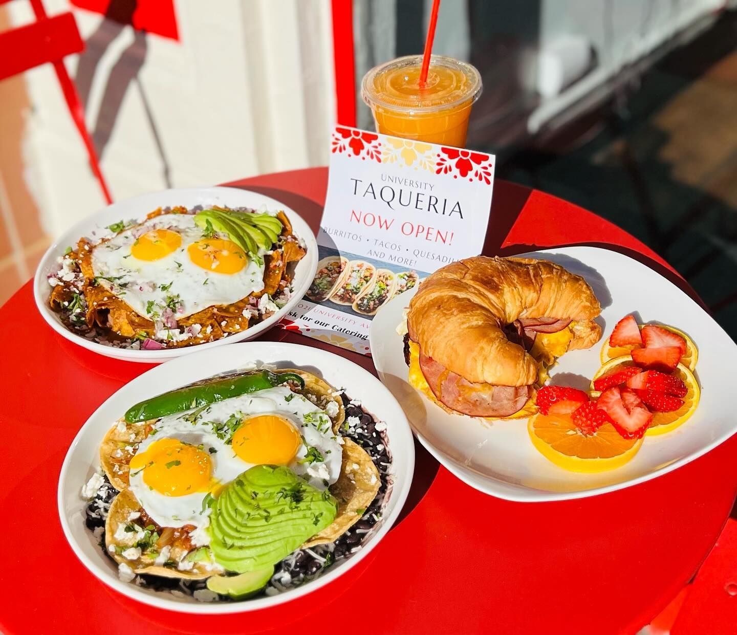 Red table with two egg-topped dishes, a croissant sandwich, fruit, and an iced drink at a café.