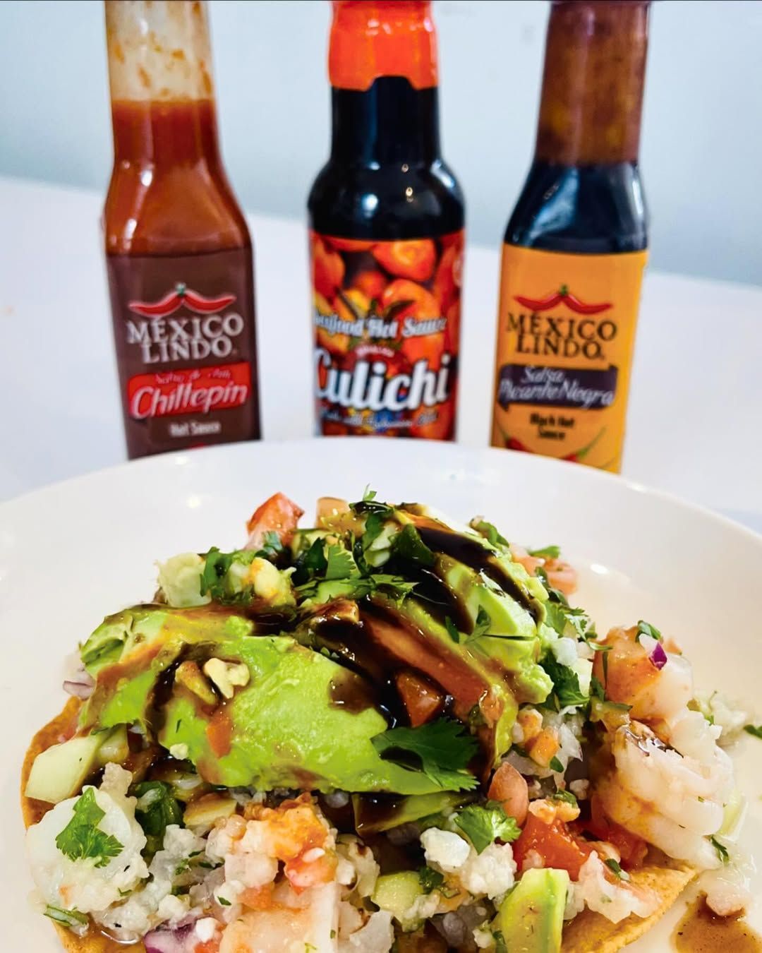 Taco bowl with avocado drizzle, rice and vegetables, with three hot sauce bottles in the background