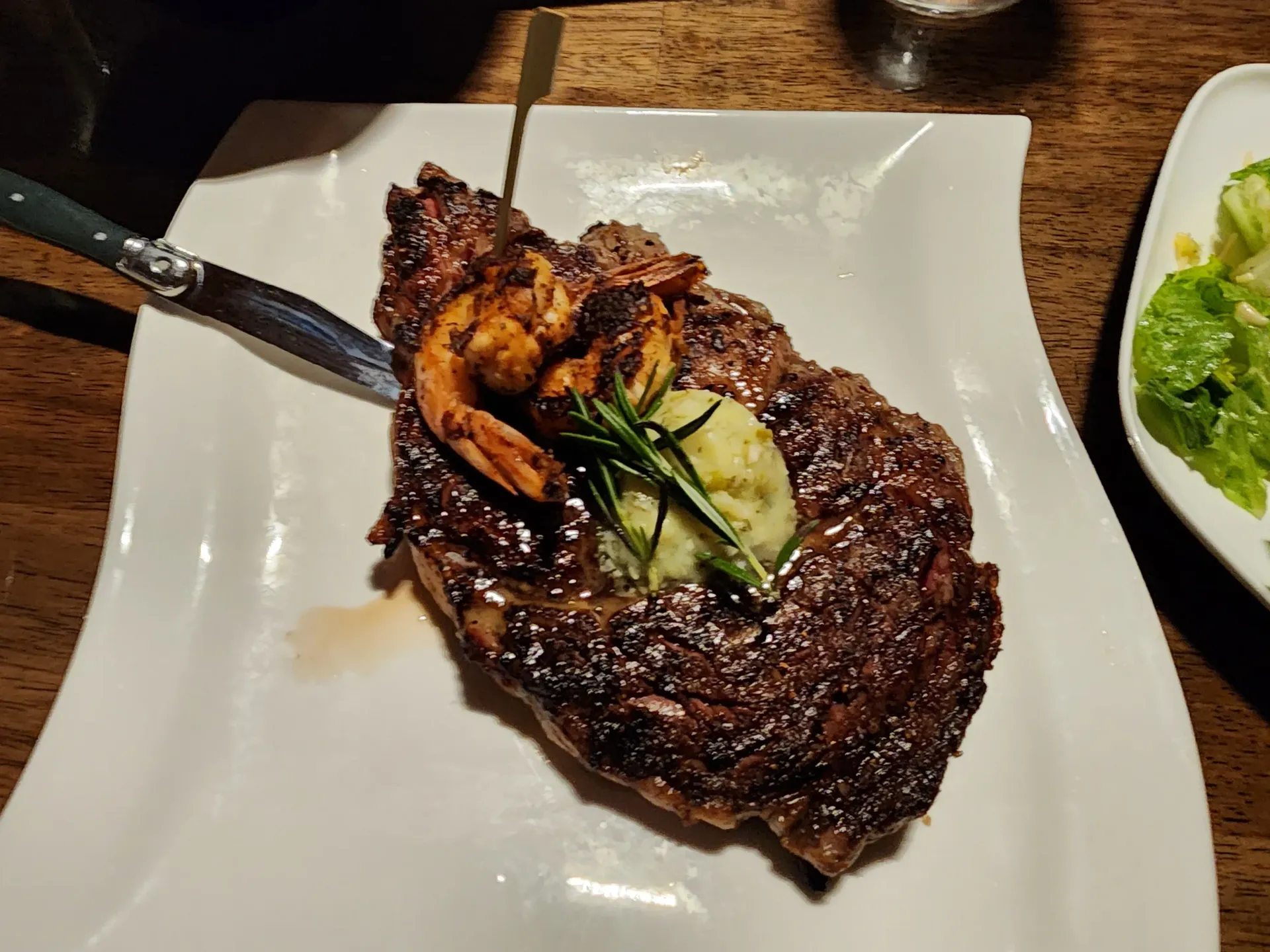 A seared ribeye steak topped with shrimp and a dollop of herb butter, served on a white plate next to a green salad.