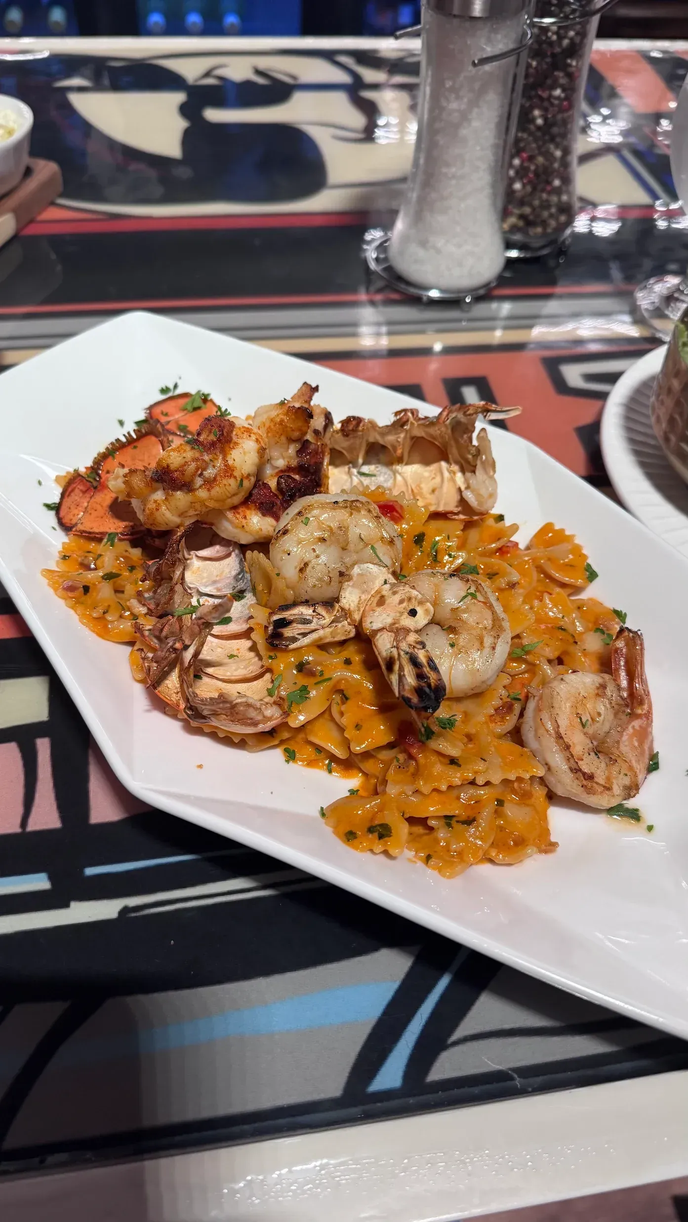 A white platter of bow-tie pasta in a creamy orange sauce topped with several grilled shrimp.